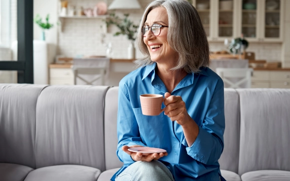 Happy senior woman with glasses sitting on a sofa and enjoying a cup of tea.