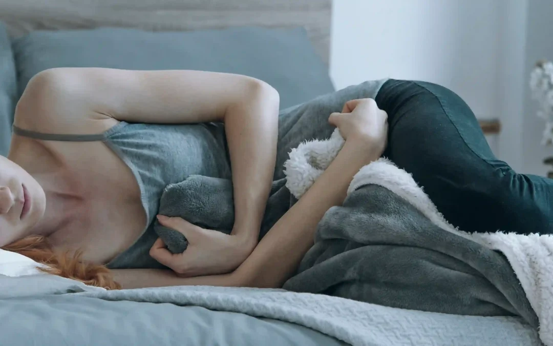 A woman lies in bed, curled up and clutching a blanket to her stomach with a pained expression.