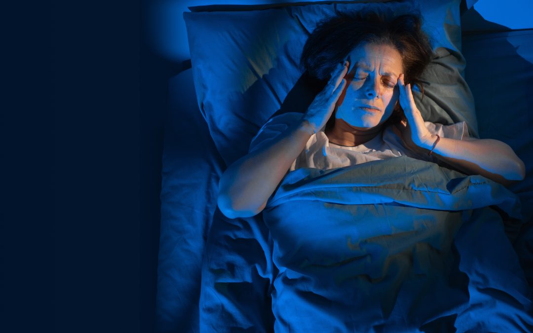 An overhead shot of a middle-aged woman in bed holding her head in pain under dim blue light.